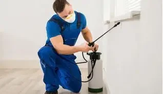 A pest control technician wearing a blue uniform and mask using a sprayer in a room.