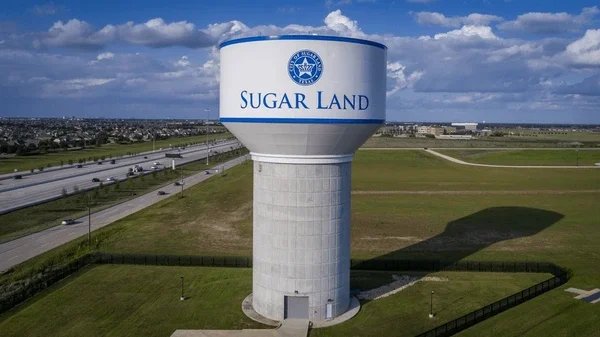 Water tower labeled 'Sugar Land' with a cityscape and highway in the background under a partly cloudy sky.