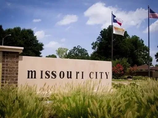 Sign reading 'Missouri City' in front of a grassy area with a Texas flag, an American flag, and a city flag flying on flagpoles under a partly cloudy sky.