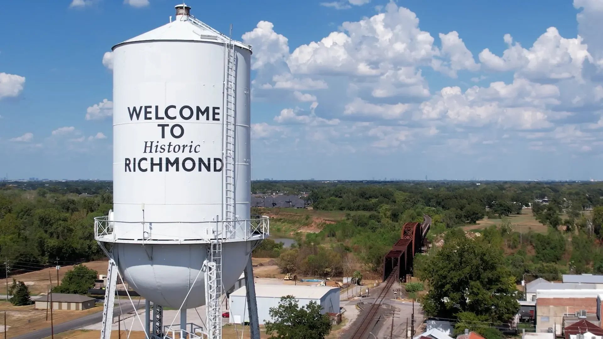 A tall white water tower with the words 'Welcome to Historic Richmond' written on it stands against a blue sky with scattered clouds. Below the tower, a landscape of trees, buildings, and railroad tracks is visible.