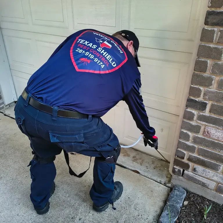A pest control technician in uniform spraying a crack in a garage door for pest treatment.