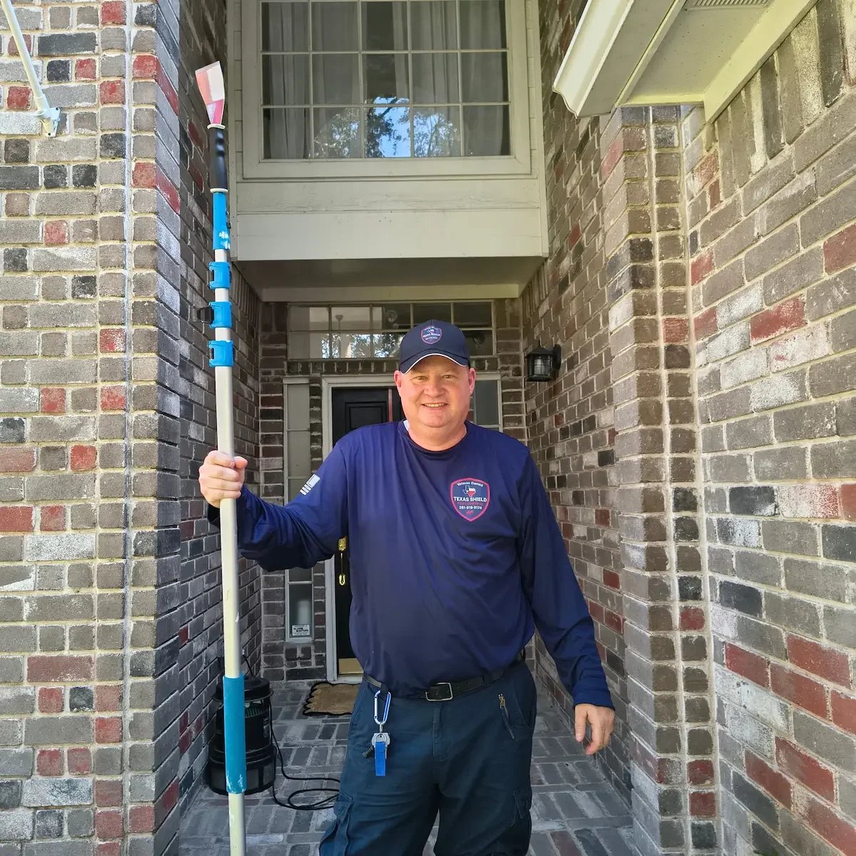 A smiling worker in a navy uniform and cap holding a pole with a brush on-top, standing in front of a brick house entrance.