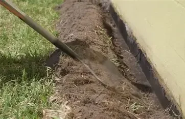 A garden hose watering a trench of soil next to a building foundation.