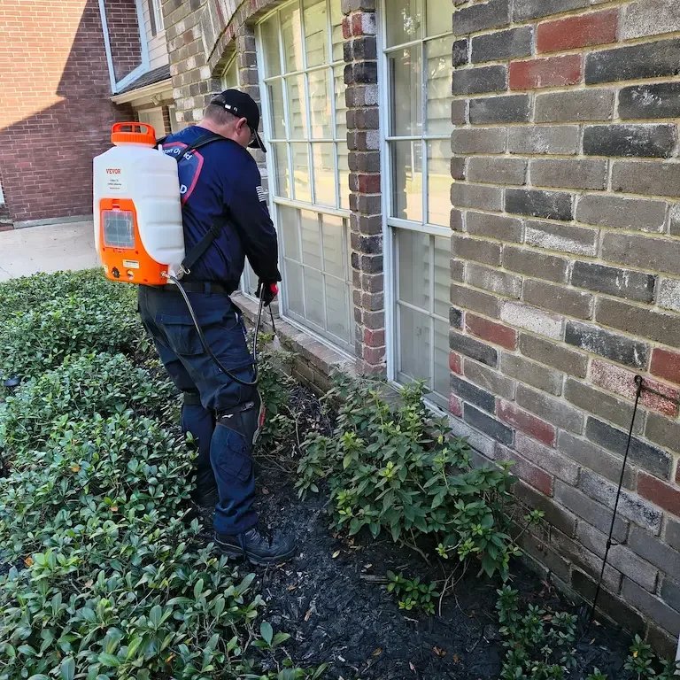 A pest control professional spraying insecticide around the foundation of a brick house.