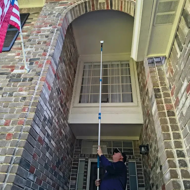 A man stands beneath a tall brick building, using an extendable pole to reach the upper windows or balcony.